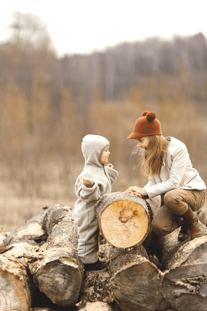 An adult and a toddler standing on a stack of logs and talking. Toddler is telling a story with arms out and hands up.